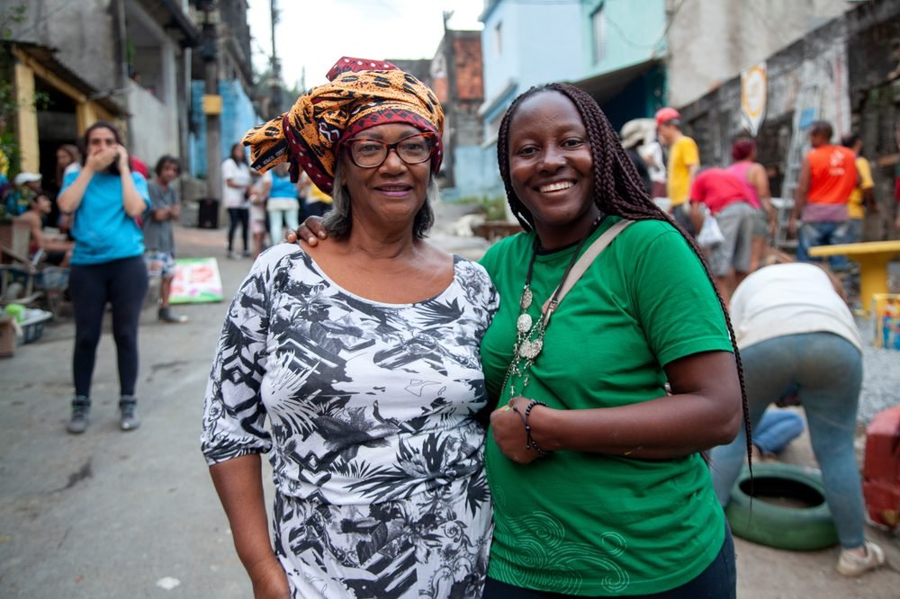 Duas mulheres lado a lado, sorrindo, em uma rua de comunidade.