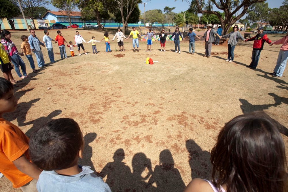 Grande roda de adultos e crianças de mãos dadas num campo de terra batida, vista por trás de duas crianças em primeiro plano.