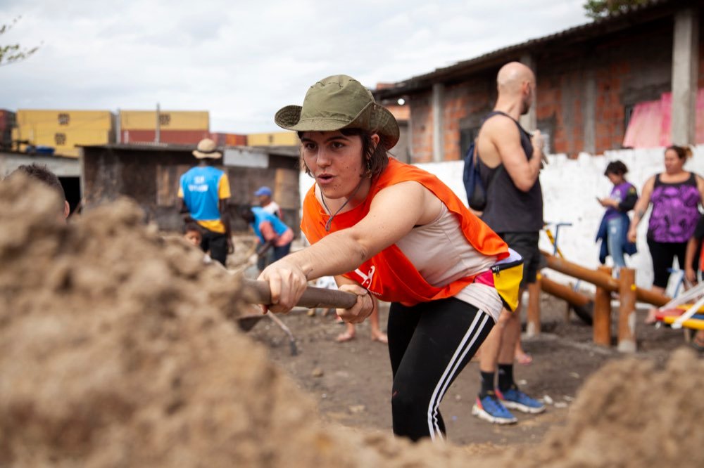 Mulher de boné e colete laranja puxando uma ferramenta de cabo de madeira sobre um monte de areia, num mutirão de rua.