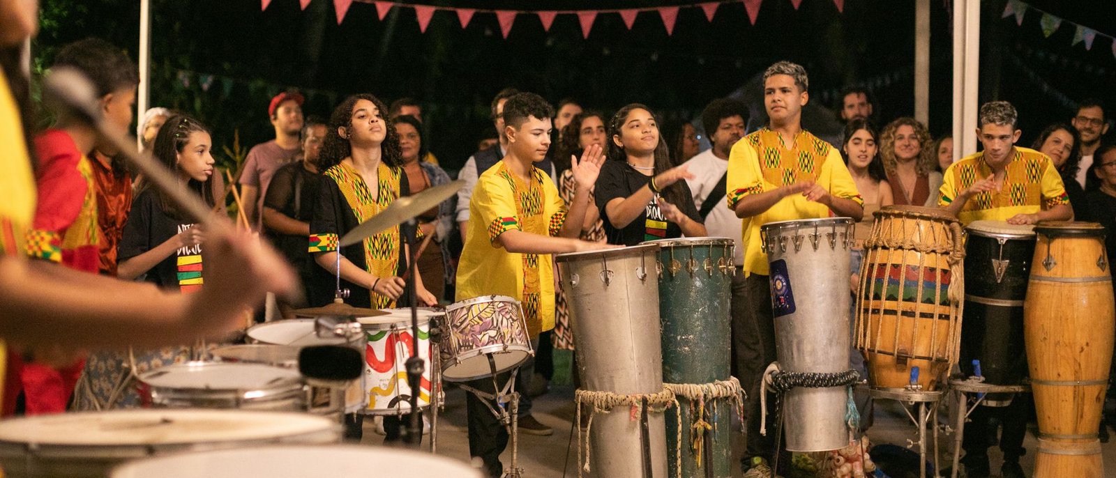 Fileira de jovens tocando tambores sob bandeirinhas, no Instituto Arte no Dique.