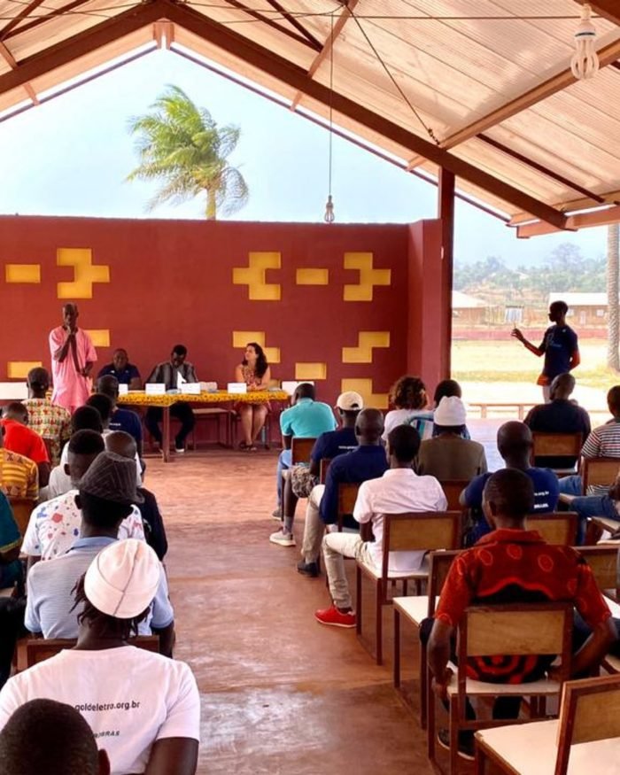 Moradores sentados em assembleia dentro do centro comunitário, voltados para a mesa de coordenação.