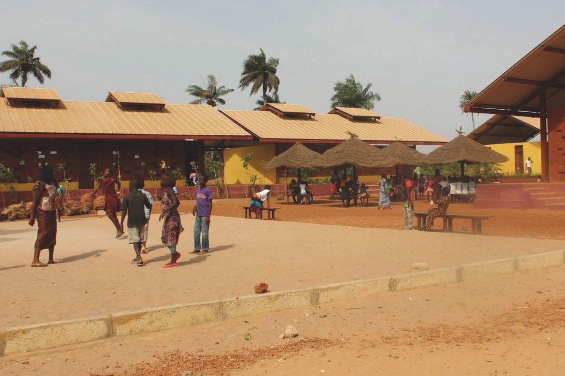Pátio de terra entre os blocos da escola comunitária, com crianças brincando.