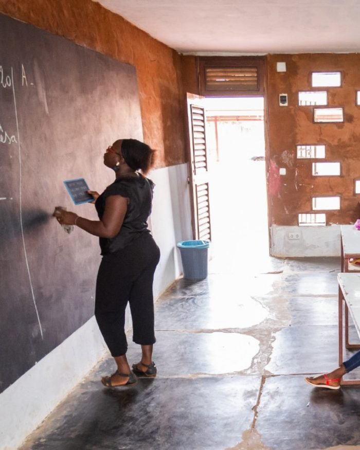 Professora escrevendo no quadro de uma sala de aula cheia de crianças, na escola construída pela comunidade.