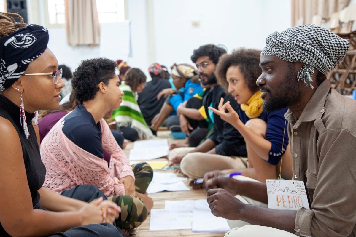 Duas fileiras de pessoas sentadas no chão, frente a frente, conversando com papéis espalhados entre elas.