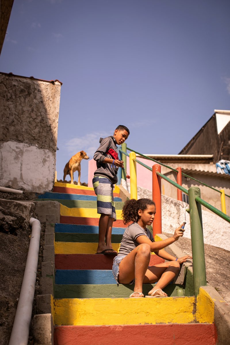Dois adolescentes pintando os degraus de uma escadaria de comunidade em faixas coloridas.