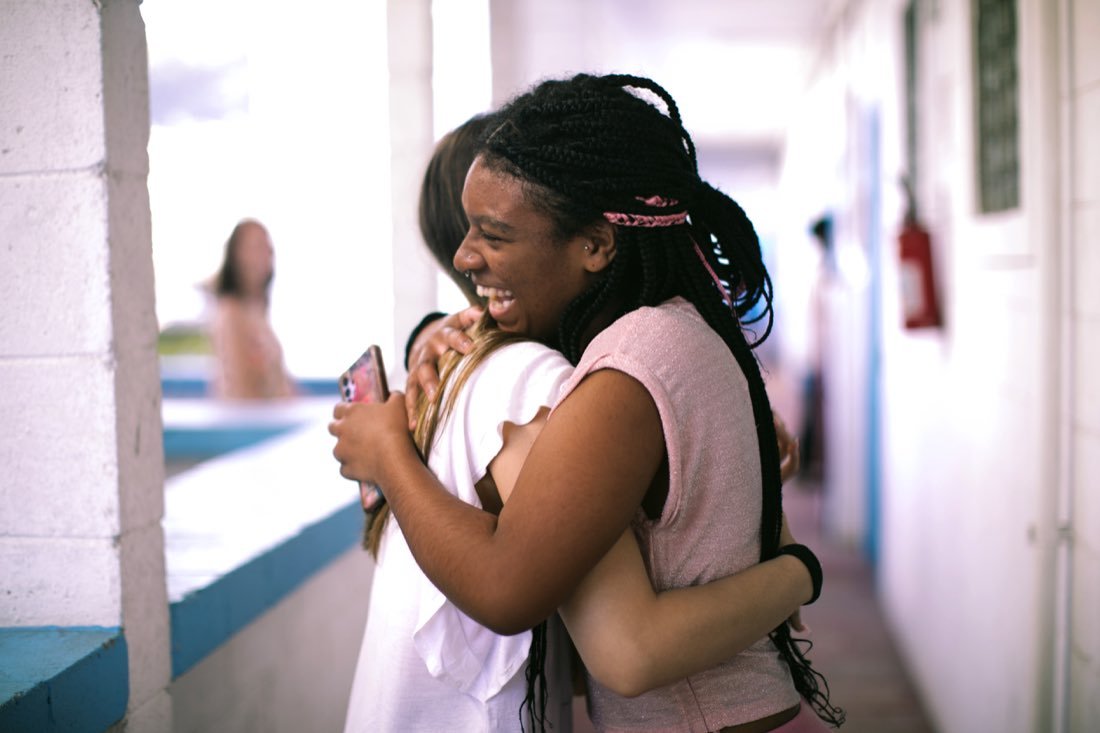 Duas adolescentes se abraçando e rindo, num corredor aberto.