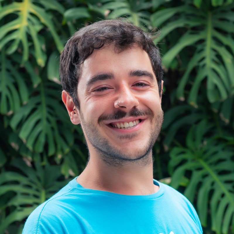 Augusto Lima Conte sorrindo, de camiseta azul, diante de uma folhagem verde.