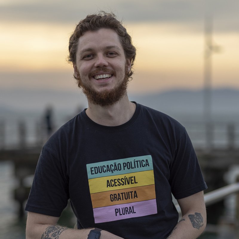 Gabriel Marmentini sorrindo ao pôr do sol, de camiseta escrita educação política acessível, gratuita e plural.