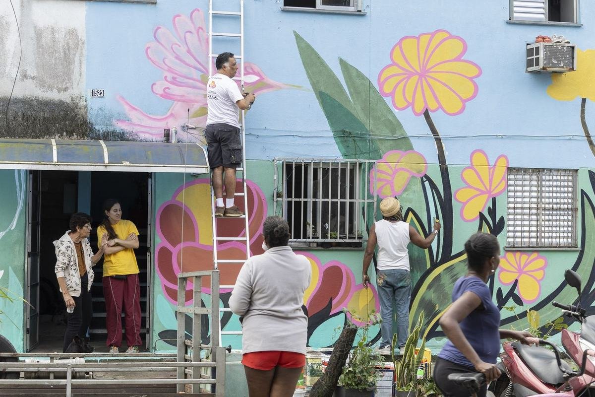 Fachada de casa sendo pintada com flores grandes, com moradores assistindo da calçada.