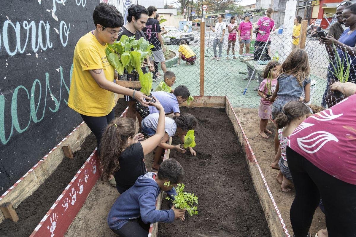 Adultos e crianças plantando juntos num canteiro montado na rua, diante de um muro escrito.