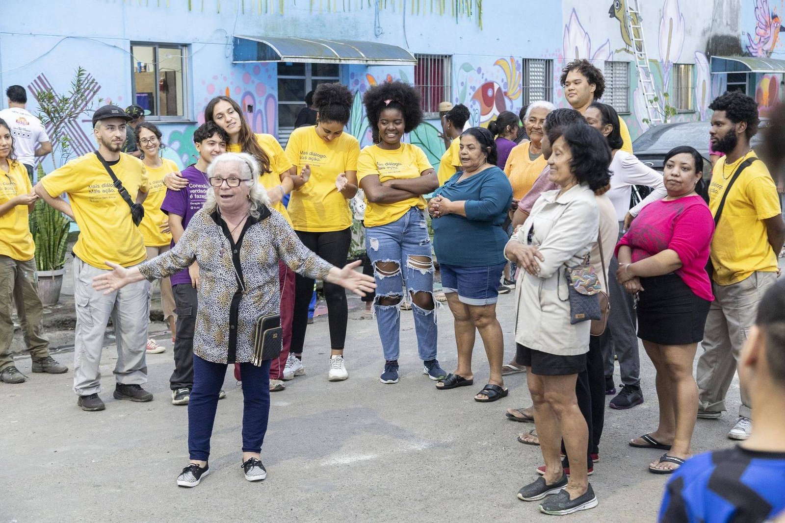 Roda de moradores e participantes de braços abertos, no meio da rua, diante de um muro pintado.