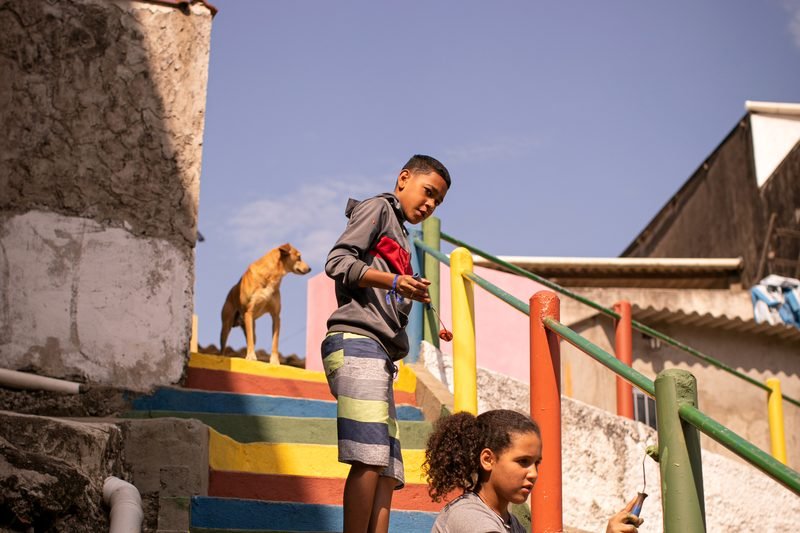 Escadaria comunitária pintada em faixas coloridas, com moradores nos degraus.