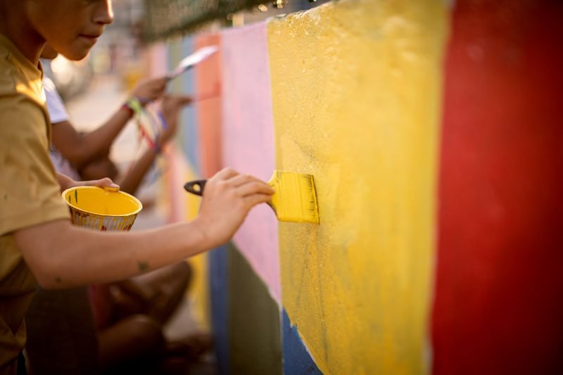 Mãos pintando um muro com rolo amarelo, ao lado de faixas vermelha e rosa.