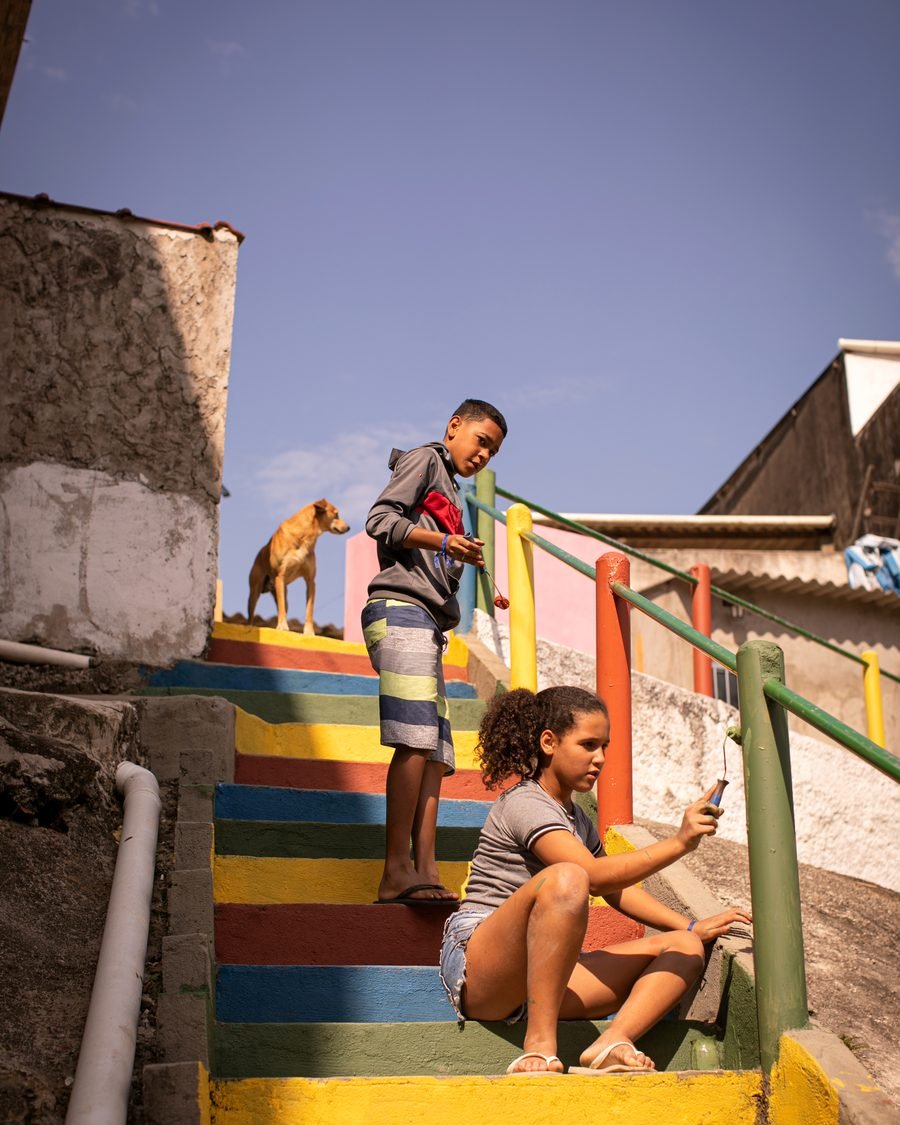 Escadaria comunitária pintada em faixas coloridas, com moradores sentados nos degraus.